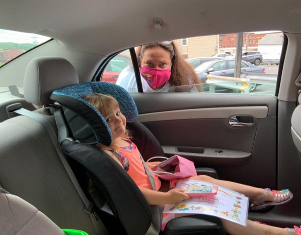 A young girl sits in a car seat, smiling and holding a colorful picture book. A woman in a pink mask leans in through the open window