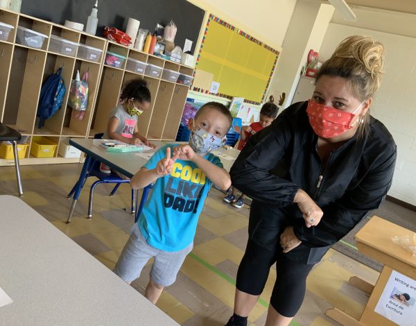 A teacher and a young boy wearing face masks smile at the camera in a classroom.
