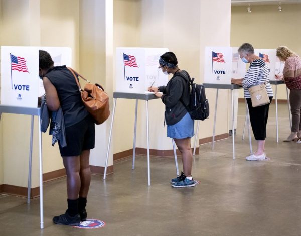 Four people are voting at separate booths in a spacious room. Each booth has an American flag and the word "VOTE" on it.