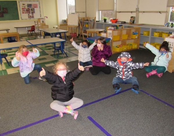 Children in colorful winter attire and masks happily pose in a classroom, thumbs up.