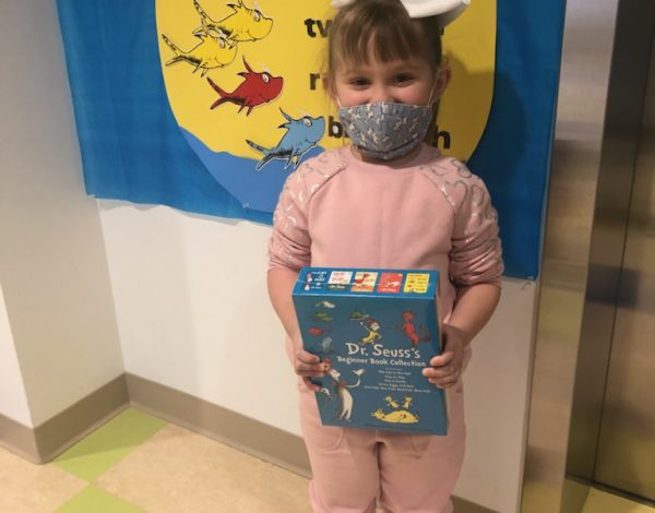 Child in pink outfit, brown boots, and mask holds Dr. Seuss book set.