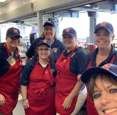 Six smiling people, wearing blue shirts, red aprons, and matching navy caps.