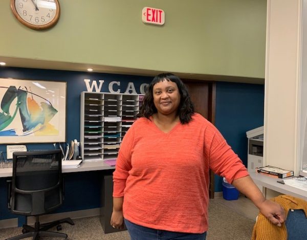 A woman in a bright orange top smiles warmly in an office setting.