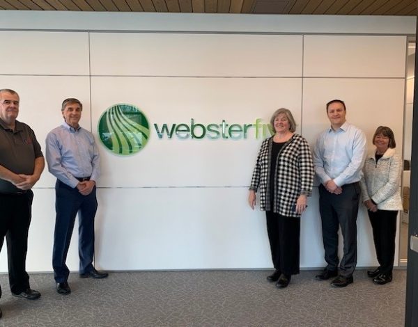 Five people stand smiling in front of a wall featuring the "websterfn" logo with a green emblem.