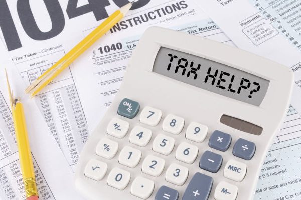 Calculator displaying "TAX HELP?" lies on a pile of 1040 tax forms and pencils.
