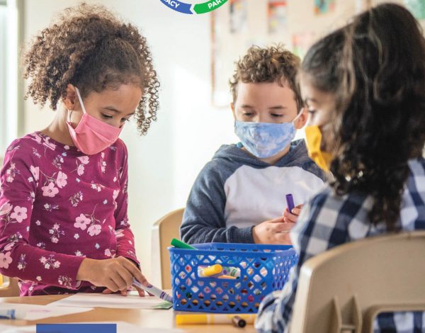 Three children sit at a classroom table wearing colorful masks, engaged in an activity.