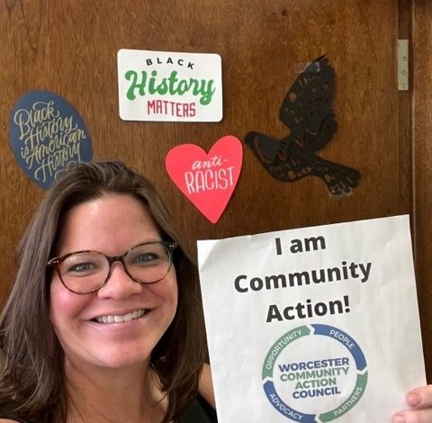 A woman smiles, holding a "I am Community Action!" sign.