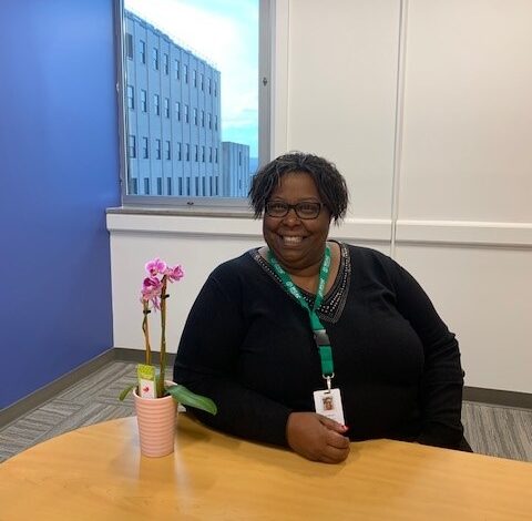 Smiling person in an office setting, wearing glasses and a green lanyard, sits at a wooden table with a pink orchid.