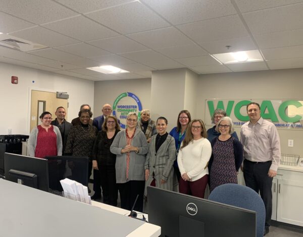 A diverse group of 13 smiling people stand in an office setting.
