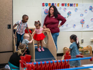 Children play on an indoor slide in a preschool classroom while two parents watch, with a colorful "Bubble Wishes" bulletin board on the wall behind them.