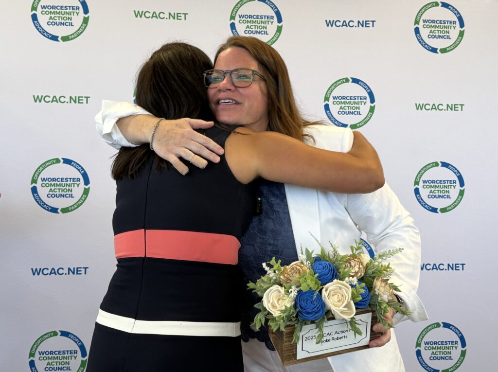 Two women embrace warmly at a Worcester Community Action Council event, one holding a floral award box labeled "2025 WCAC Action Hero, Brooke Roberts."