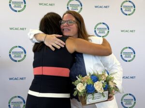 Two women embrace warmly at a Worcester Community Action Council event, one holding a floral award box labeled "2025 WCAC Action Hero, Brooke Roberts."