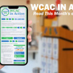 A hand holds up a smartphone displaying the "WCAC in Action" monthly eNewsletter, with sections on Fuel Assistance, Early Education & Care, and Resiliency visible on screen. To the right, bold white text reads "WCAC in Action: Read This Month's eNewsletter!" against a blurred background of a community office space with a wooden podium.