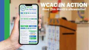 A hand holds up a smartphone displaying the "WCAC in Action" monthly eNewsletter, with sections on Fuel Assistance, Early Education & Care, and Resiliency visible on screen. To the right, bold white text reads "WCAC in Action: Read This Month's eNewsletter!" against a blurred background of a community office space with a wooden podium.