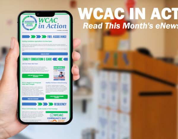 A hand holds up a smartphone displaying the "WCAC in Action" monthly eNewsletter, with sections on Fuel Assistance, Early Education & Care, and Resiliency visible on screen. To the right, bold white text reads "WCAC in Action: Read This Month's eNewsletter!" against a blurred background of a community office space with a wooden podium.